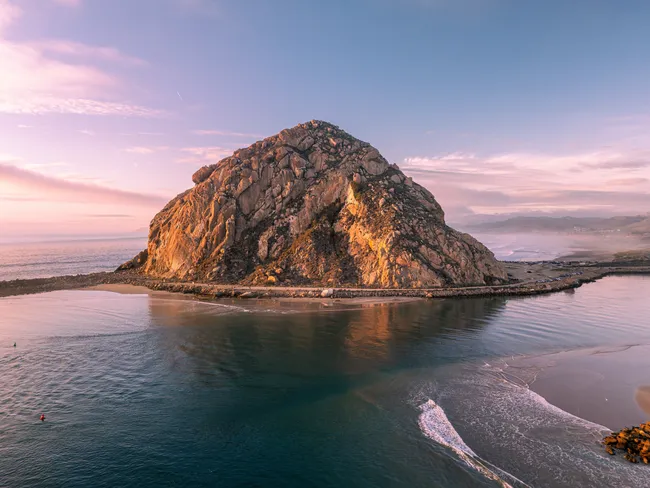 Morro Rock rises from calm coastal waters at sunset, its rugged granite face glowing in soft pink and golden light while gentle waves curve along the shoreline beneath a pastel sky.