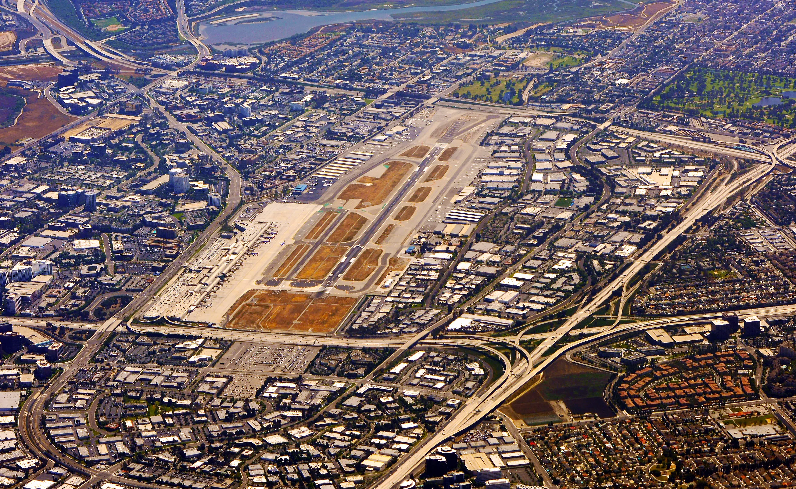 A wide, high-altitude aerial shot of an airport with parallel runways and white terminals, tightly bordered by a dense grid of commercial buildings and suburban housing. Multiple major highways with complex interchanges curve around the perimeter of the airfield, and a small blue body of water is visible in the distance under a hazy sky.