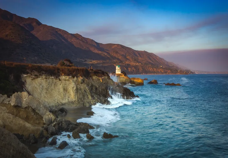 A high-angle, wide landscape shot of a rocky coastline at dusk. Jagged, tan-colored cliffs drop into a deep blue ocean with white surf crashing against the rocks and a small hidden beach. In the mid-ground, a white lifeguard tower with a teal top stands on a rocky point. Massive, dark coastal mountains rise in the background under a sky with soft orange and blue sunset tones.