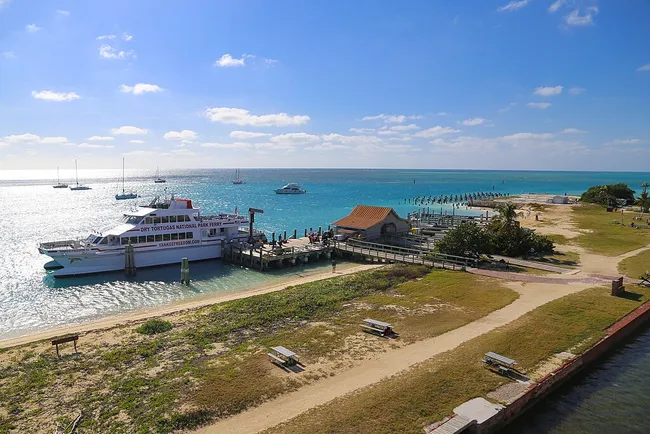 A ferry docked at Dry Tortugas National Park with turquoise waters, sailboats in the distance, and a sunny blue sky.