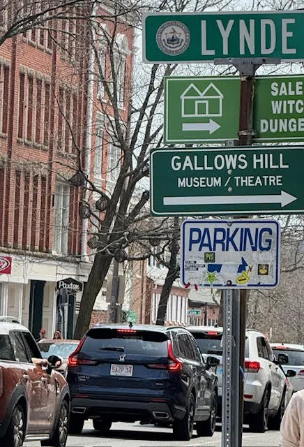 Street view on Lynde Street in Salem, featuring directional signs for Gallows Hill Museum, Salem Witch Dungeon, and a parking area, with red-brick buildings and cars lining the road.