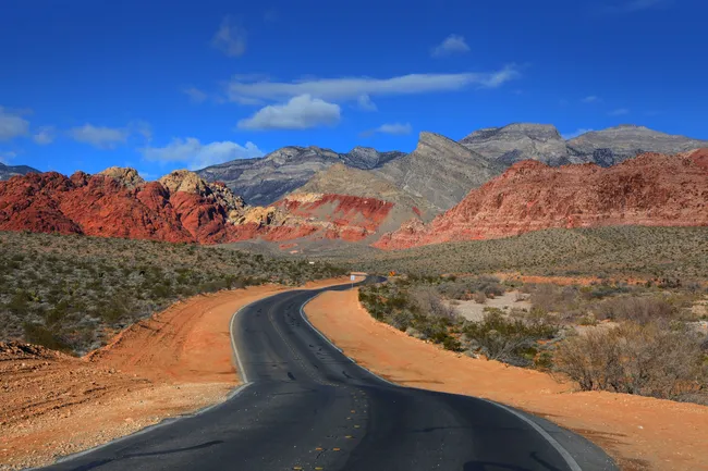 Curving desert highway leading toward red rock formations and layered mountains under a clear blue sky.