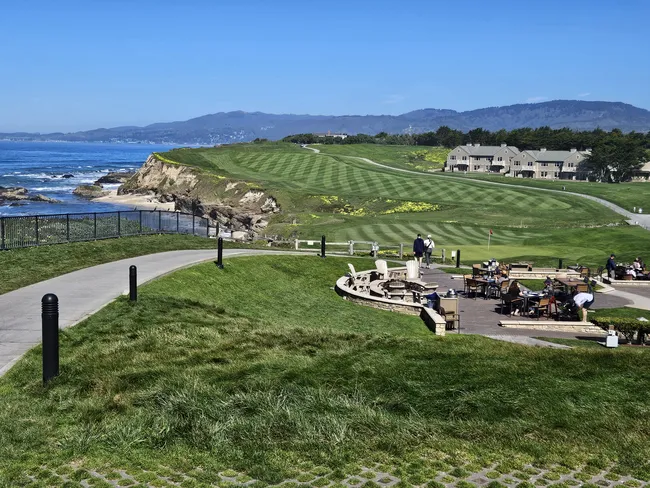 A manicured coastal golf course stretches along dramatic oceanfront cliffs, with rolling green fairways overlooking the shoreline. A walking path winds through the landscape, while people gather on an outdoor terrace with panoramic views of the coast and distant hills.