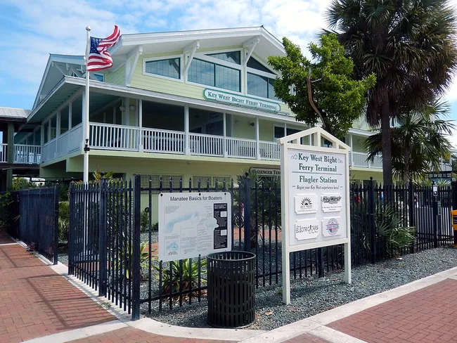Key West Bight Ferry Terminal with a U.S. flag, informational signs, and palm trees surrounding the entrance.