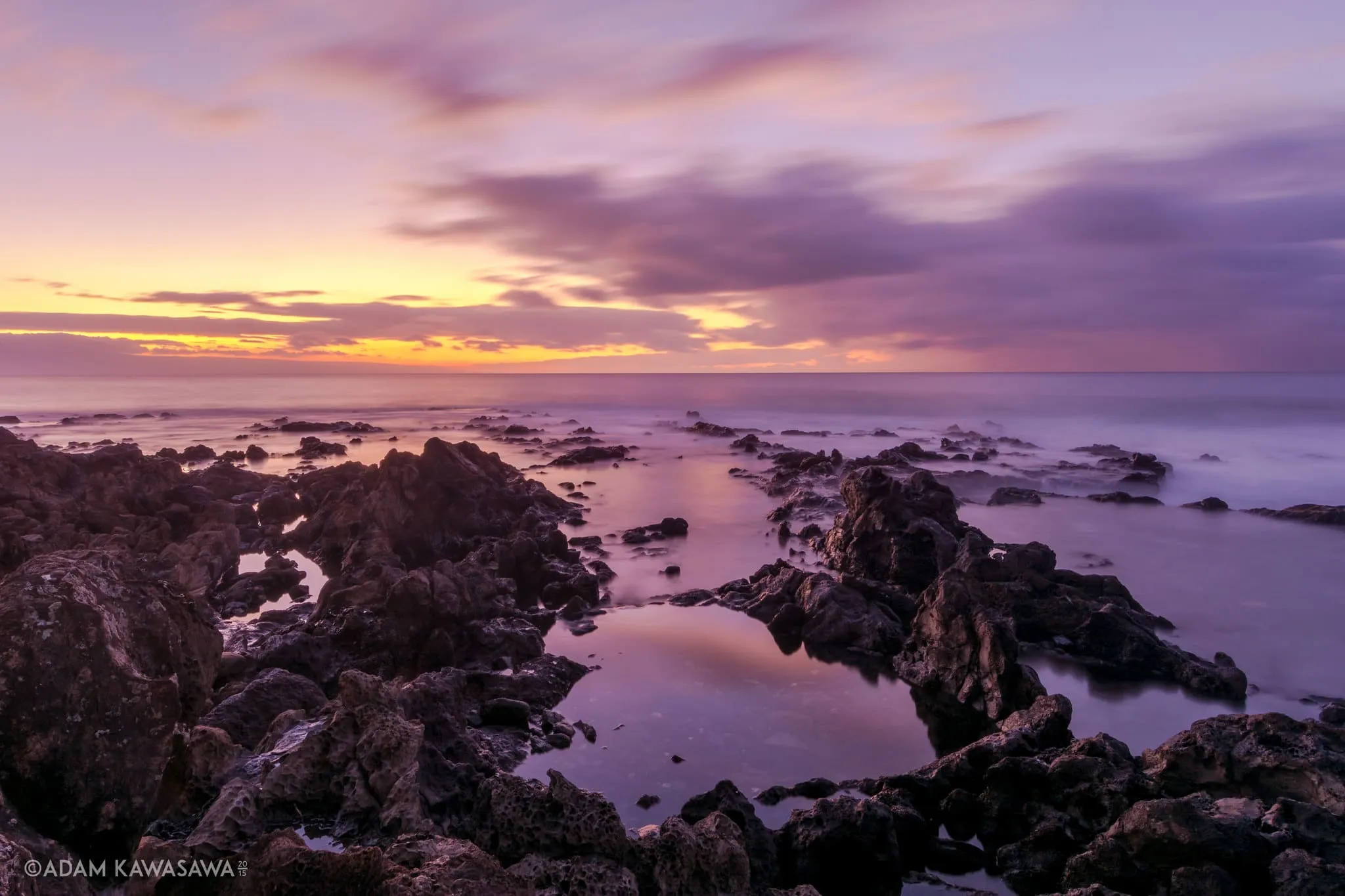 Rocky shoreline at sunset with purple clouds and tide pools reflecting the fading light.