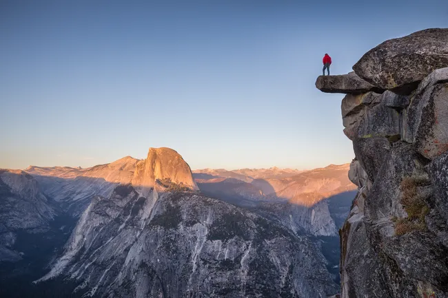 A person wearing a bright red jacket stands on the tip of a flat, overhanging rock ledge extending high over a deep mountain valley. In the background, a massive, rounded granite peak is partially illuminated by the warm, golden light of a setting sun against a clear, pale sky.