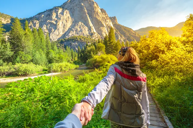 A first-person view of a person holding another person's hand while walking along a wooden boardwalk in a lush green meadow. In the background, a large, light-grey granite mountain rises above a dense forest of evergreen trees under a bright sky.