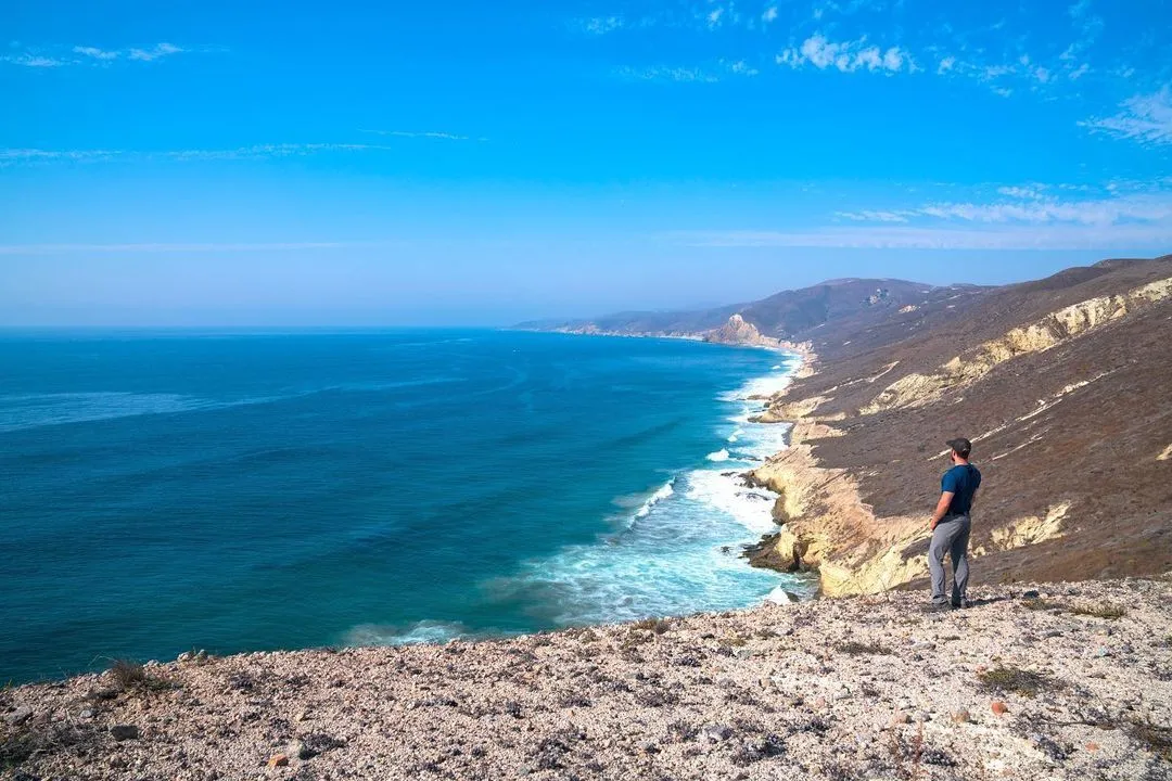 A person stands on a rugged coastal cliff overlooking a vast blue ocean, with waves crashing against steep shoreline bluffs that stretch into the distance beneath a clear sky.