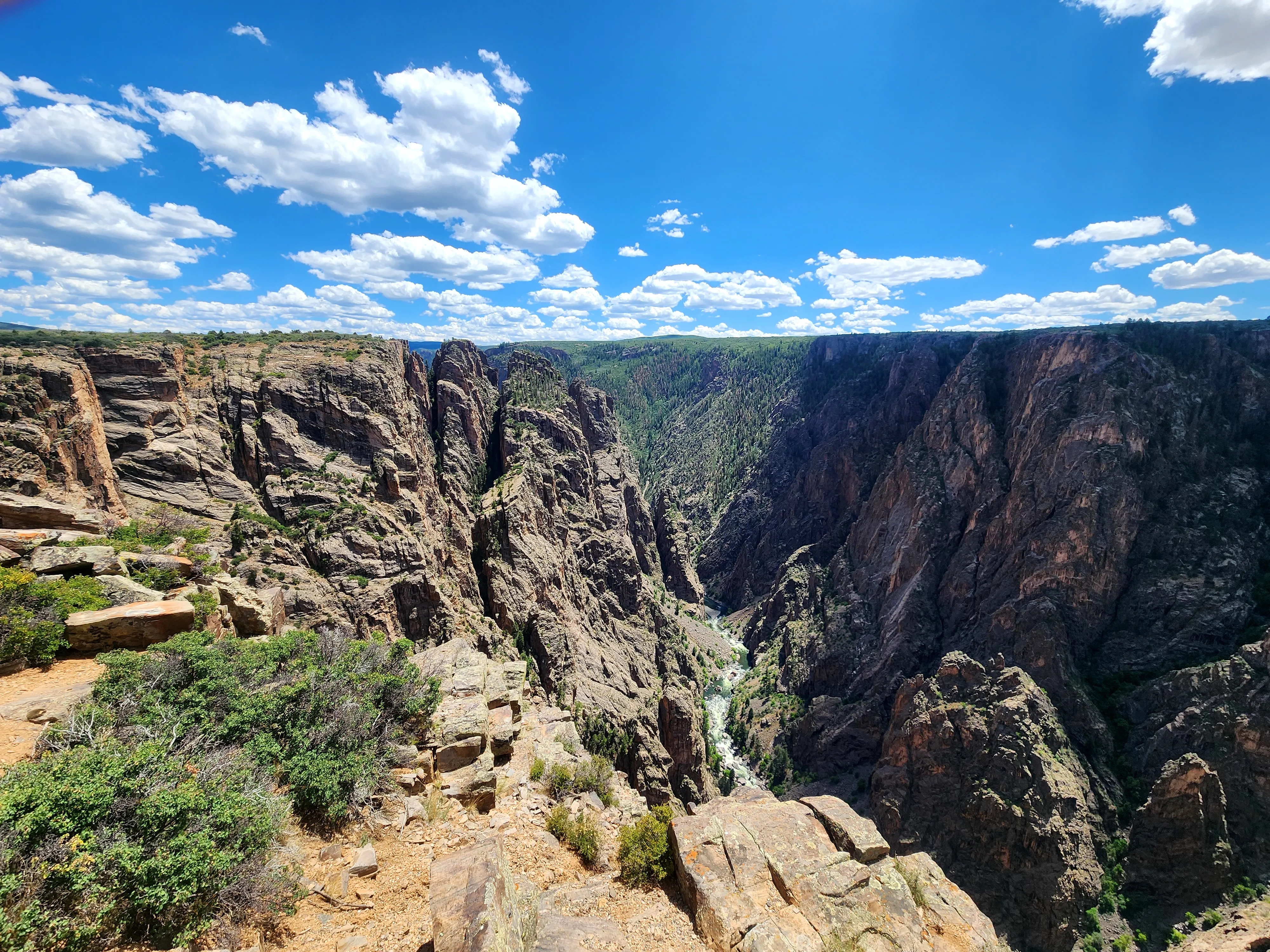 A dramatic view of Black Canyon’s steep, jagged cliffs dropping into a narrow gorge with a river below under a bright blue sky filled with scattered clouds.