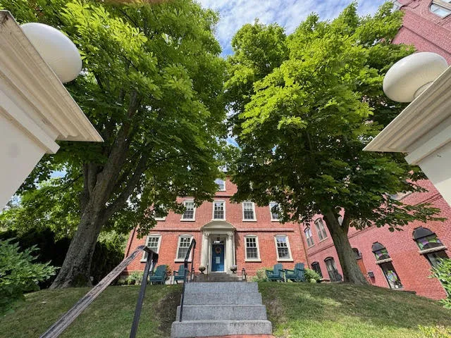 Red-brick Merchant Hotel framed by tall green trees, viewed from bottom of stairway lined with white posts.