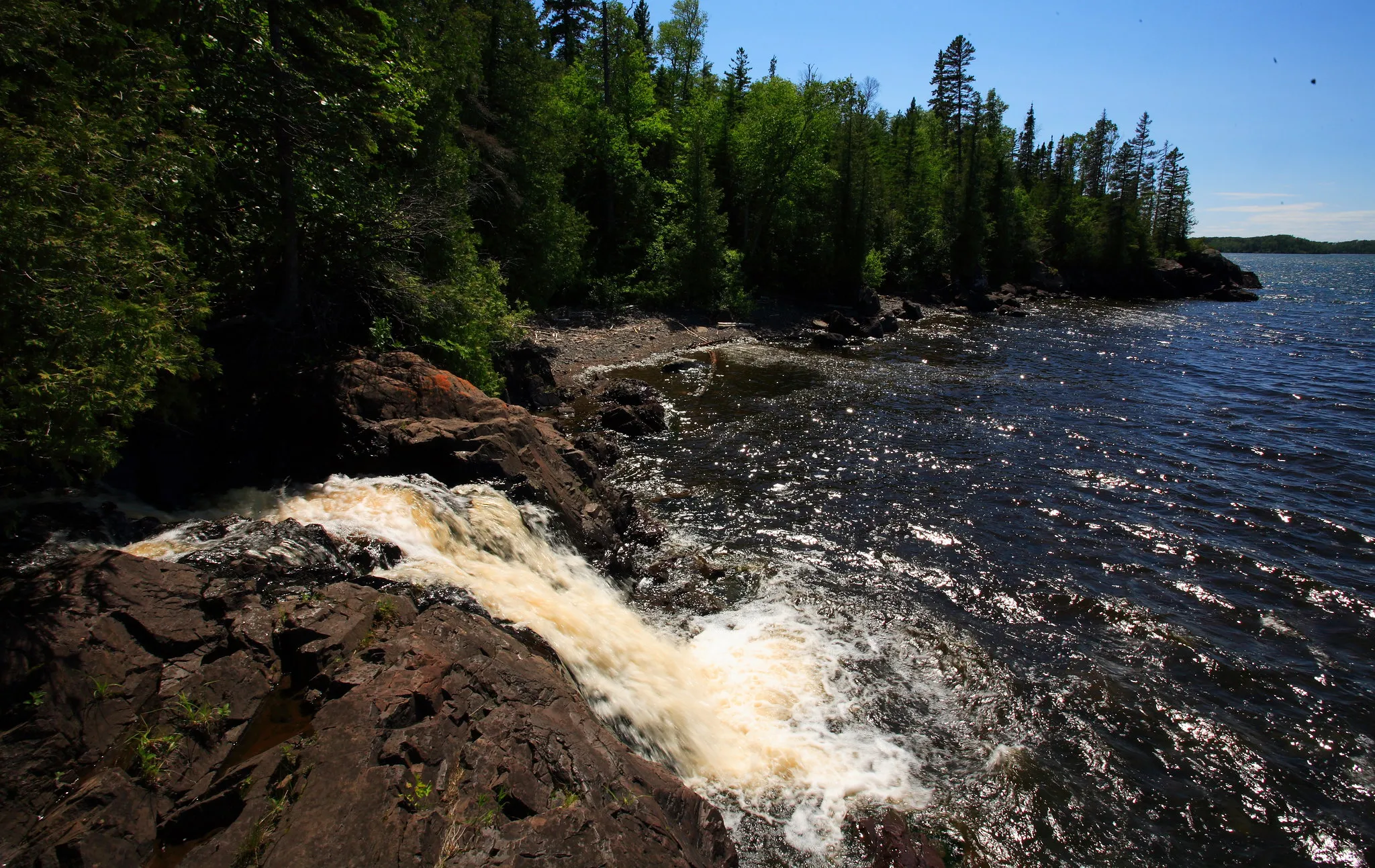 Alt text: Small waterfall cascades over dark rocky cliffs into a large lake, bordered by a dense evergreen forest along a rugged shoreline under clear, sunny skies.