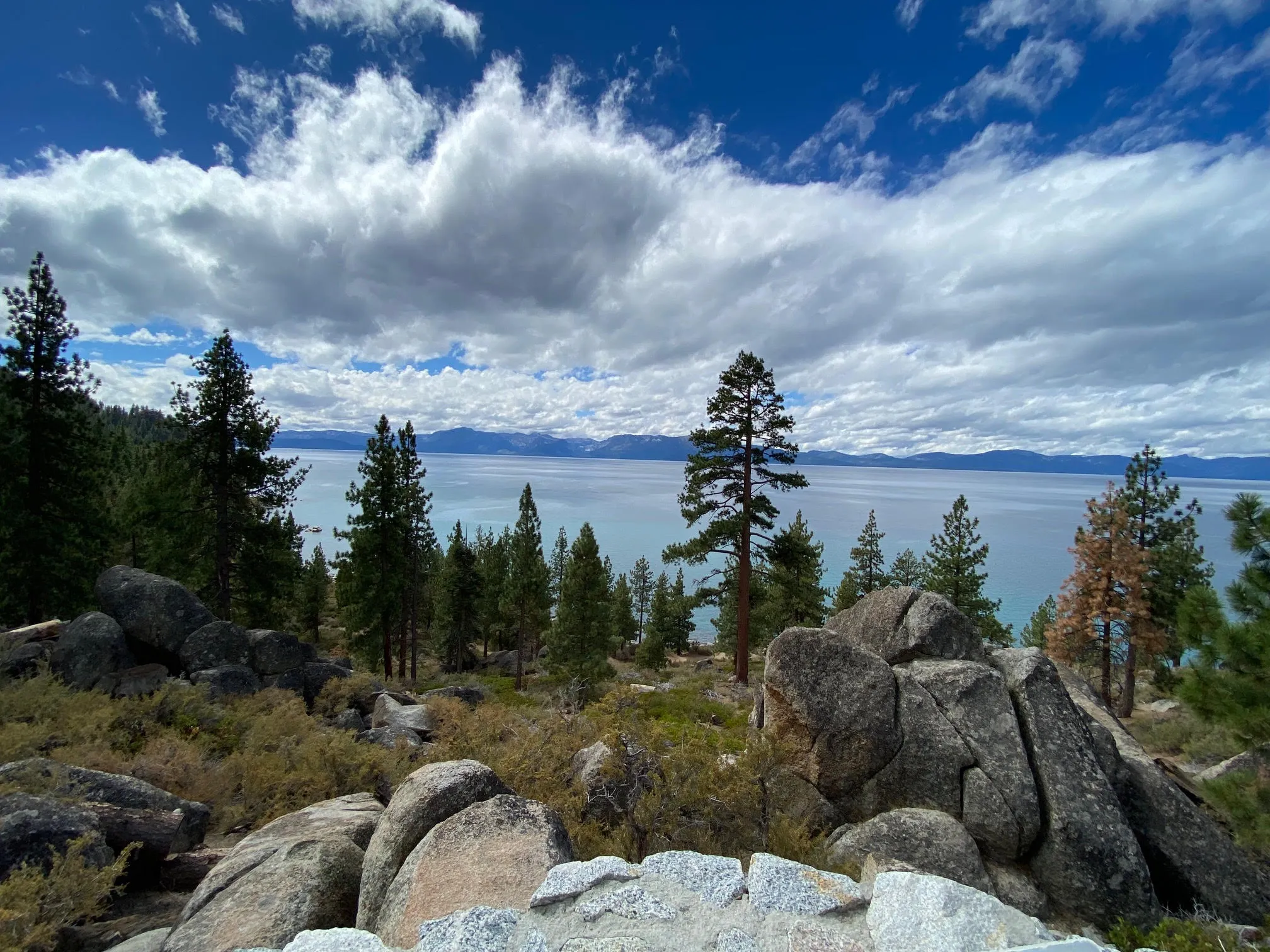 Scenic viewpoint at Lake Tahoe with boulders, pine trees, turquoise water, and dramatic cloudy sky.
