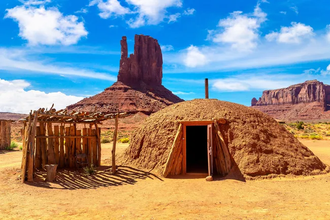 Traditional Navajo hogan and wooden shelter in a desert landscape with towering red rock formations.