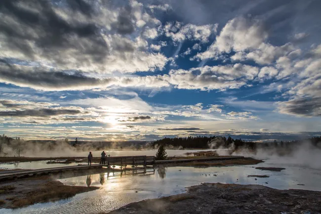 Sunset over a steaming Yellowstone boardwalk with visitors reflected in geothermal pools.