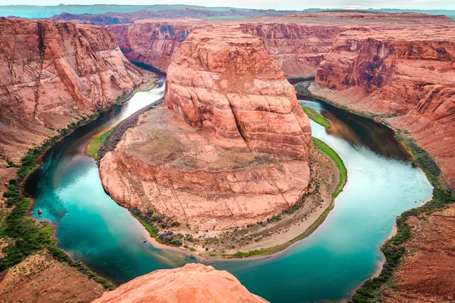 An aerial view of Horseshoe Bend near Page, Arizona, where the Colorado River makes a dramatic 270-degree horseshoe-shaped meander around a massive sandstone butte. The vivid turquoise-green river wraps tightly around the towering rust-red and orange layered rock formation, with sparse desert vegetation and a small sandy beach visible at the river's edge far below. Sheer canyon walls and a vast plateau stretch toward the horizon under an overcast sky.