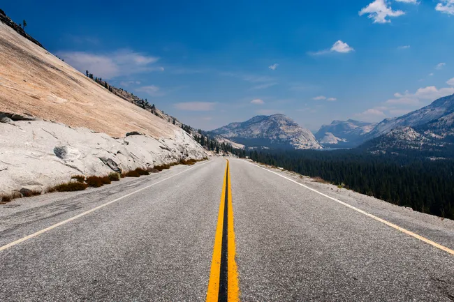 A long, paved two-lane highway with a bright double yellow center line stretches toward distant, jagged mountains under a clear blue sky. A massive, smooth light-colored granite slope rises sharply on the left side of the road, while a dense green evergreen forest fills the valley on the right.