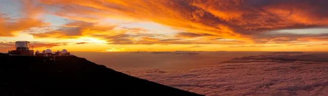 Sunrise above a sea of clouds, with an observatory silhouetted on a mountain ridge under glowing orange skies.