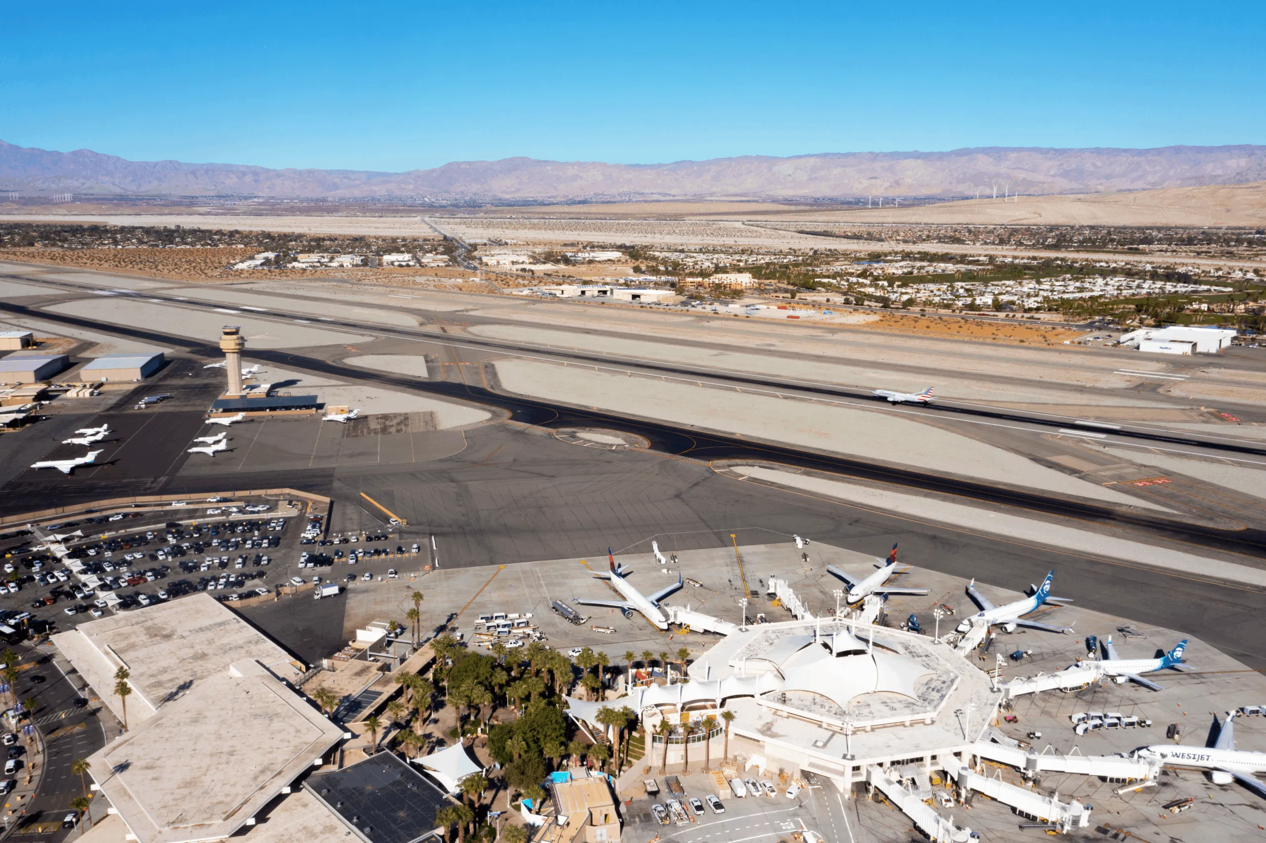 A high-angle aerial view of a desert airport terminal with a distinctive white, tent-like roof structure. Several commercial airplanes are parked at gates or taxiing on the vast grey runways. In the background, a flat desert landscape is bordered by a range of hazy, light-brown mountains under a clear blue sky.
