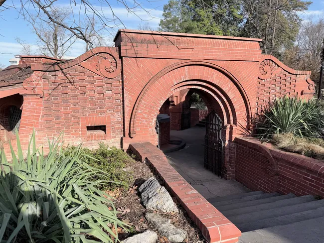 A decorative red brick gateway structure featuring a large ornate rounded arch with concentric brick moldings and scrollwork details on either side of the parapet. A black wrought iron gate is set within the arch, leading to a brick-paved walkway beyond. Low plantings of yucca and shrubs flank the structure on both sides. Bare and evergreen trees are visible in the background under a partly cloudy blue sky. The architectural style suggests a historic garden entrance, likely on the grounds of the Smithsonian Institution in Washington, D.C.