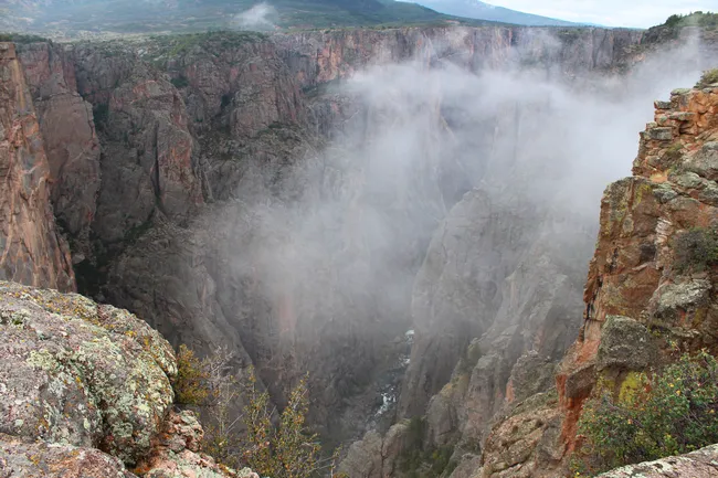 Fog drifts through the deep chasm of Black Canyon of the Gunnison, partially obscuring the rocky cliffs and river below in a moody, dramatic scene.