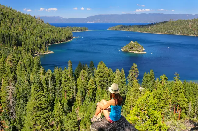 Woman sitting on a rock overlooking a deep blue lake surrounded by dense pine forest and distant mountains.