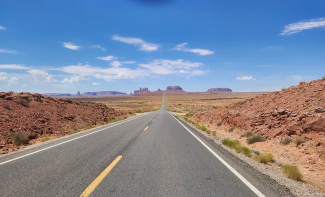 Straight highway stretching into the vast desert landscape of Monument Valley under a bright blue sky.