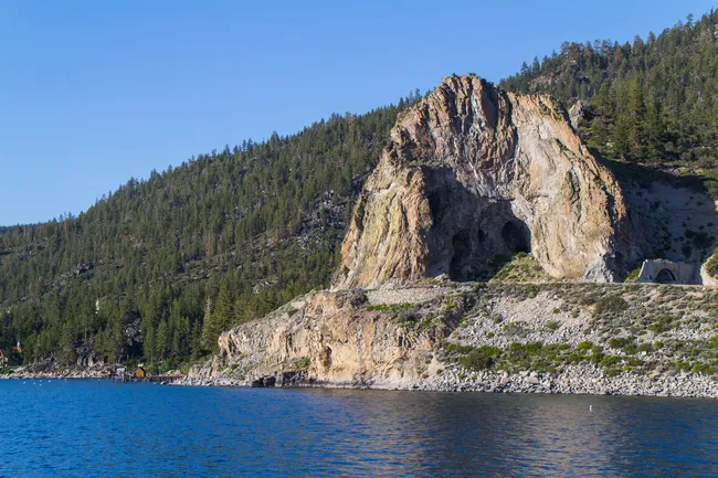 Rocky cliff with a large cave opening rising above a clear blue lake and forested hillside under a bright sky.