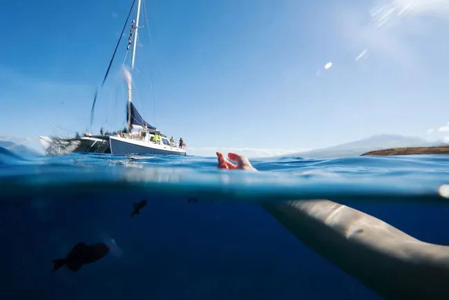 Split-level view showing a snorkeler’s arm underwater with a sailboat floating above in clear blue ocean.