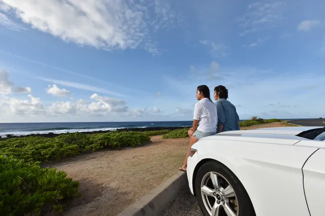 Two people sit on the hood of a white car parked near the ocean, gazing at the waves and enjoying a scenic coastal view under a blue sky with scattered clouds.