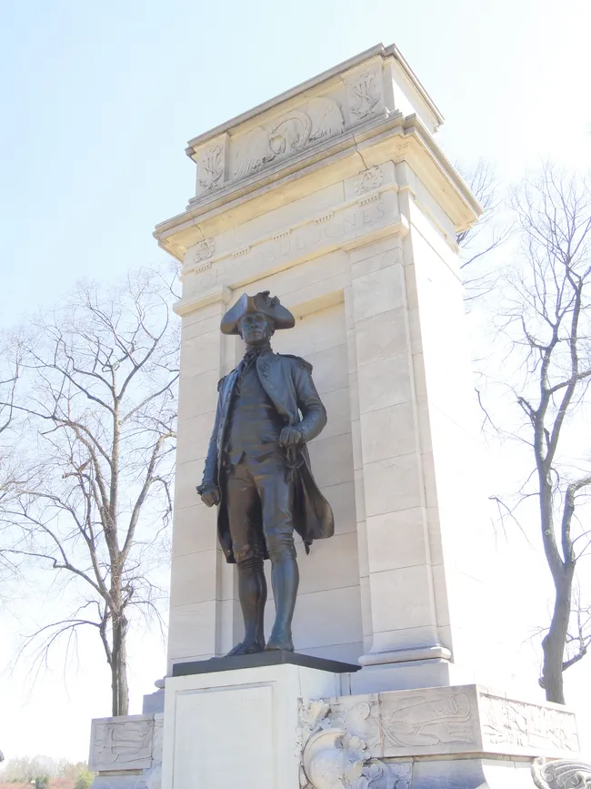 A low-angle view of the John Paul Jones Memorial in Washington, D.C. A bronze statue of John Paul Jones, the American Revolutionary War naval hero, stands in full 18th-century naval uniform — tricorn hat, long coat, and breeches — mounted against a tall rectangular limestone pedestal with decorative relief carvings near the top. Bare winter trees are visible on either side against a bright, hazy sky.