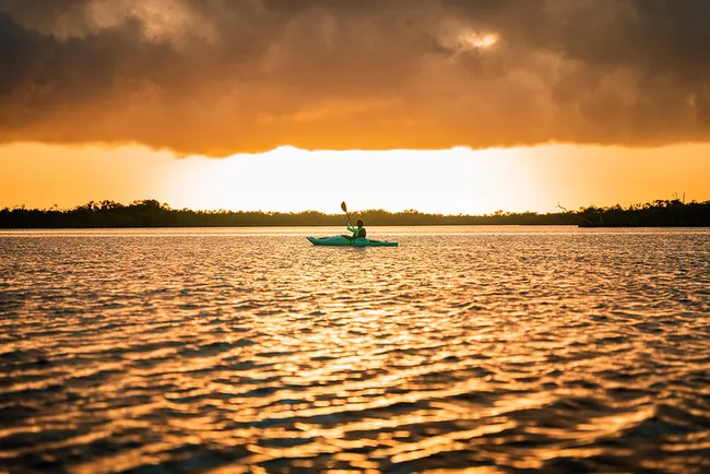 A lone kayaker paddles across golden waters at sunset in Everglades National Park under dramatic clouds.