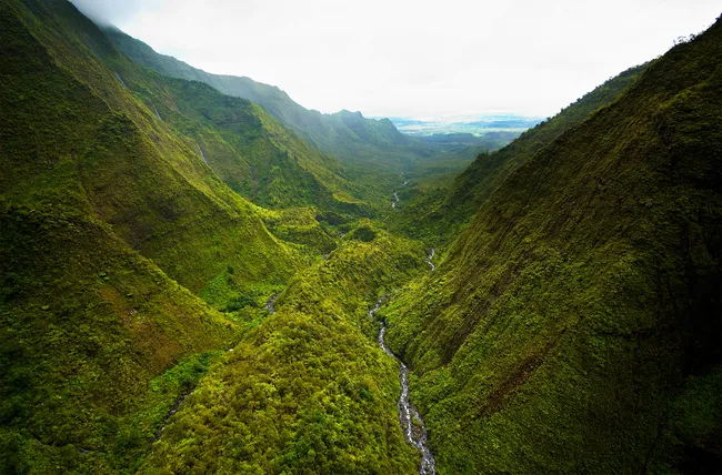 A lush, green valley in Hawaii stretches into the distance, with a narrow river winding through steep mountain slopes.