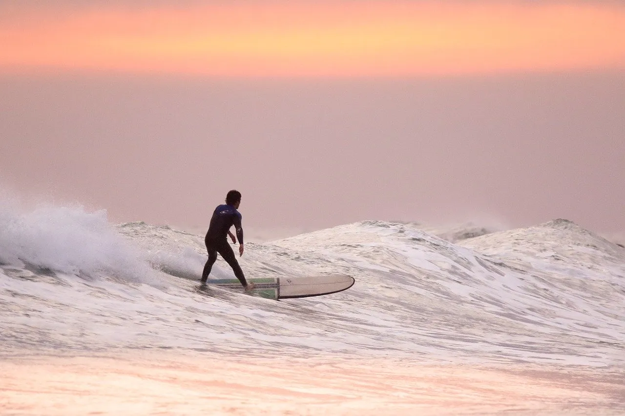 Surfer riding a gentle wave at sunset, with soft orange and pink hues lighting the sky.