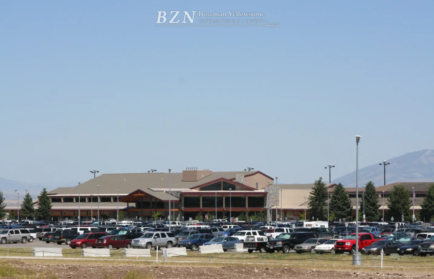 Bozeman Yellowstone International Airport exterior view with a large parking lot full of cars and mountain backdrop under clear blue skies.