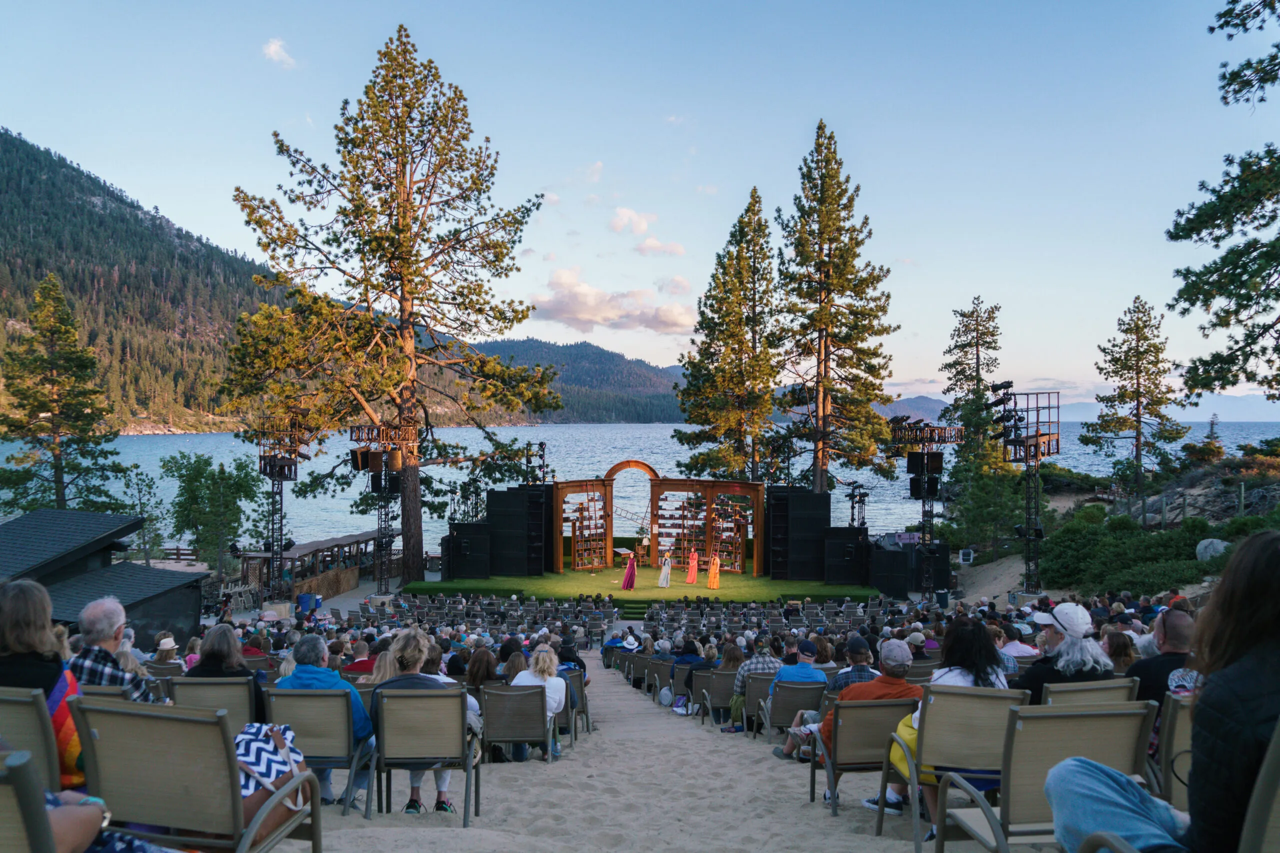 Outdoor lakeside theater at Lake Tahoe with audience seated on sandy slope watching a stage surrounded by pine trees and mountains.