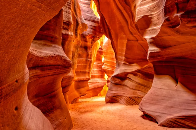 An eye-level perspective from deep inside a narrow slot canyon. The undulating sandstone walls are a vibrant mix of fiery orange and deep red, with horizontal sediment lines visible in the rock. Warm, bright light spills through a narrow vertical opening in the distance, illuminating the fine, sandy canyon floor. The smooth, flowing shapes of the walls create a sense of movement and depth.