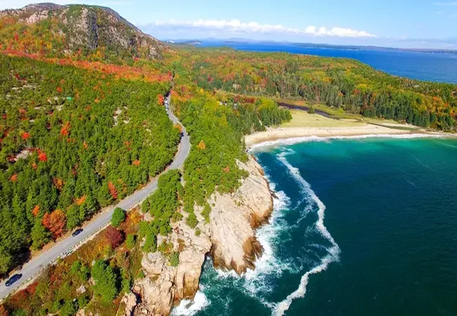 Aerial view of Acadia National Park’s coastline showing fall foliage, rocky cliffs, and turquoise ocean waves.