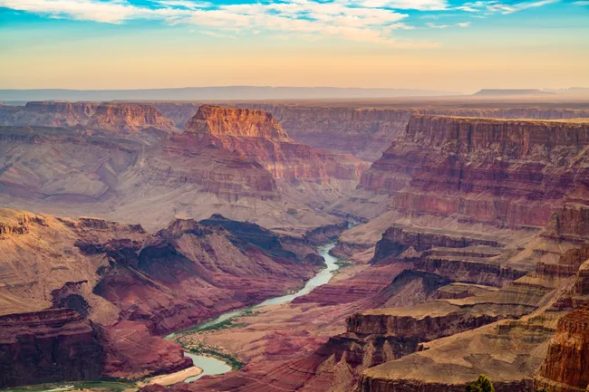 A horizontal, high-perspective landscape of the Grand Canyon’s Inner Gorge. The green Colorado River snakes through the bottom of the canyon, bordered by steep, dark red and brown rock walls. In the mid-ground, massive sandstone buttes and plateaus rise with distinct horizontal banding, illuminated by soft golden afternoon light. The far horizon shows a flat desert plateau under a blue sky with thin, wispy white clouds.