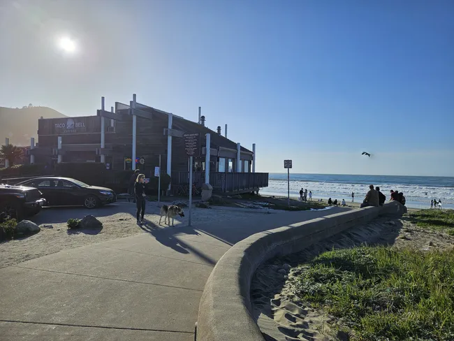 A beachfront restaurant sits beside a sandy shoreline under a bright, clear sky, with ocean waves rolling in the background. People walk and gather along the beach and seawall, while a person stands on the sidewalk with two dogs near parked cars.