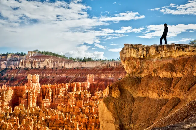 A lone silhouetted figure stands on the edge of a rocky cliff at Sunset Point in Bryce Canyon National Park, Utah, gazing out over a vast amphitheater filled with thousands of vivid orange, red, and cream-colored hoodoo spires. The densely packed eroded rock formations stretch across the canyon floor and up layered canyon walls, topped by a forested plateau of dark green pines in the background. The scene is set under a dramatic blue sky with sweeping white clouds.