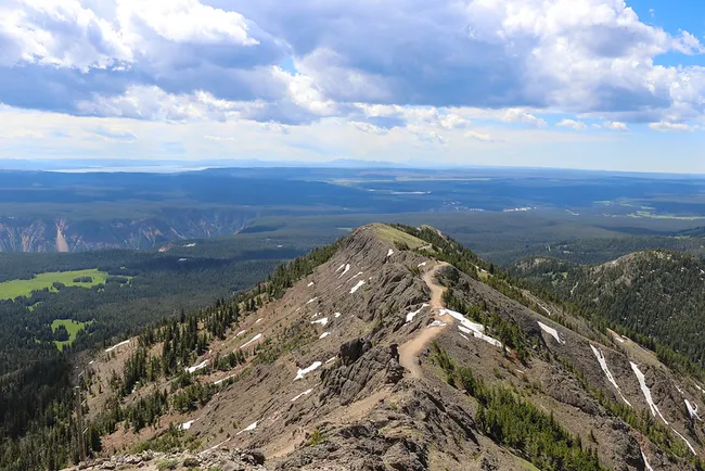 Expansive view from the summit of Mount Washburn in Yellowstone National Park, showing a rugged ridgeline trail with patches of snow and panoramic forested valleys below.