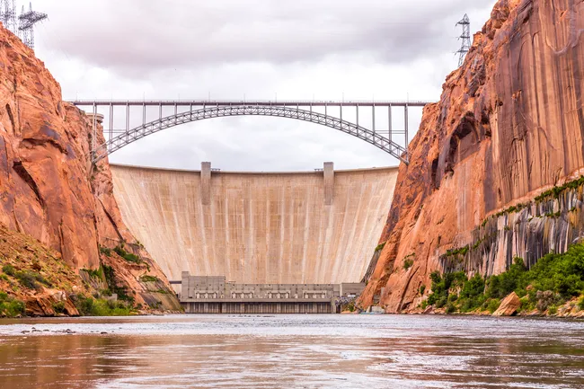 A low-angle, eye-level shot from the surface of the Colorado River looking toward the massive Glen Canyon Dam. The high, vertical red sandstone canyon walls frame the river on both sides. A large, steel arch bridge (Glen Canyon Bridge) spans the canyon in front of the smooth, curved concrete face of the dam. The water in the foreground is calm and brown, reflecting the overcast gray sky above. Large power lines are visible on the canyon rims above the dam.
