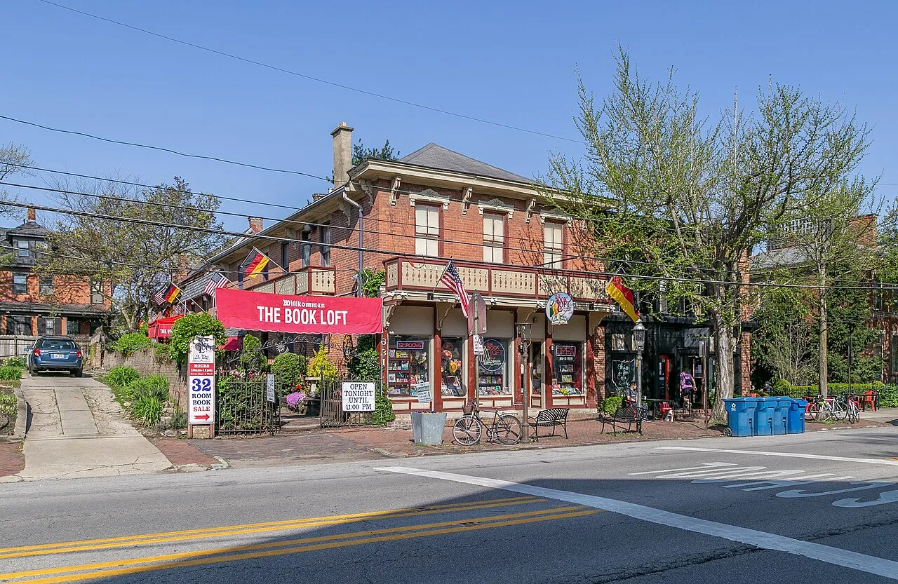 Street-level view of The Book Loft, an independent bookstore housed in a two-story red brick building on a corner lot in German Village, Columbus, Ohio. A large red banner across the front reads "Willkommen — The Book Loft." Signs near the entrance advertise "32 Room Book Sale" and "Open Tonight Until 11:00 PM." American and German flags decorate the balcony railing. A bicycle is parked out front alongside iron benches, potted plants, and decorative ironwork fencing. Bare spring trees frame the building under a clear blue sky.