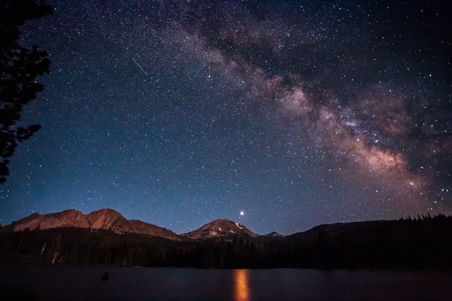 A wide-angle night shot of a dark mountain range silhouetted against a deep blue sky filled with thousands of stars and the glowing, purple-and-orange band of the Milky Way galaxy. A calm lake in the foreground reflects a single bright celestial body.