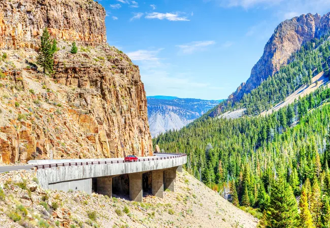 A red car drives along a raised mountain roadway built against a steep rocky cliff, overlooking a forested valley and distant ridges under a bright blue sky.