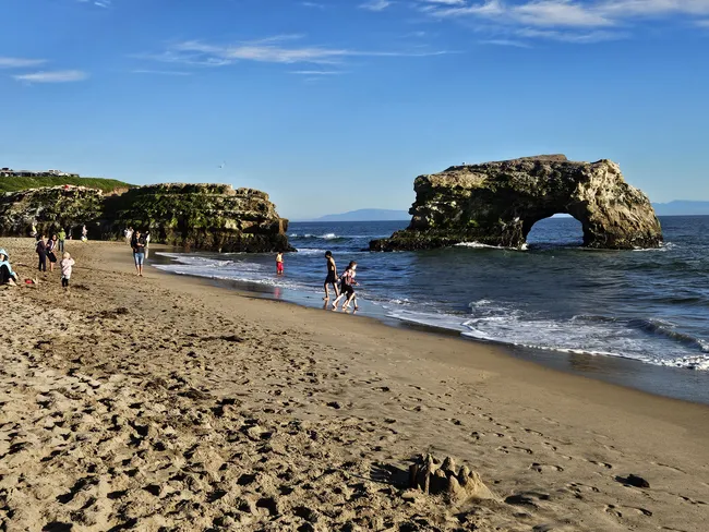 A sandy beach curves along the shoreline where visitors walk, play, and explore the water’s edge on a sunny day. Just offshore, a large natural rock arch rises from the ocean, creating a striking coastal landmark against the blue sky.