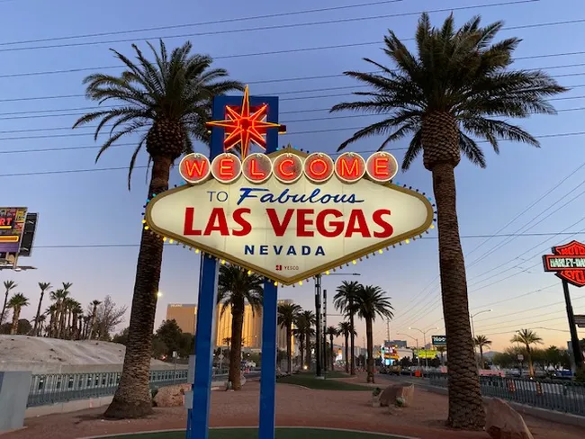 Illuminated roadside sign with palm trees and city lights in the background during dusk.