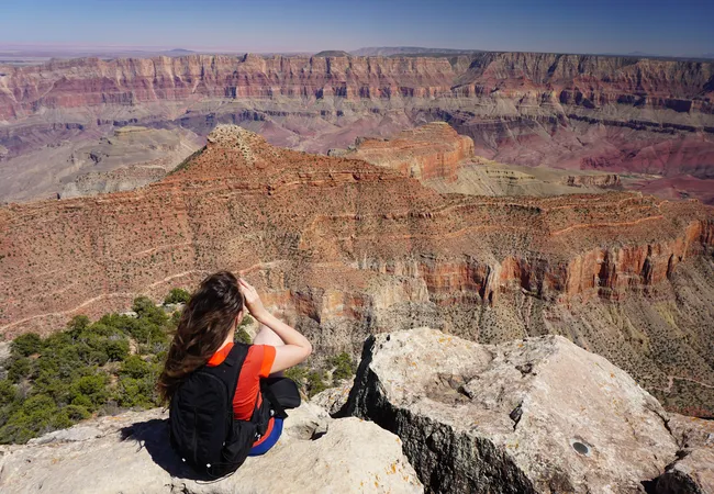A person with long brown hair, wearing a red shirt and a black backpack, sits on a light-colored rocky ledge with their back to the camera. They are looking out over a vast, deep canyon filled with massive red and orange layered rock formations stretching to a flat horizon under a clear sky.