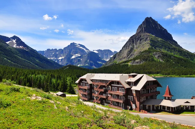 Many Glacier Hotel sits beside a lake with dramatic mountain peaks rising behind it in Glacier National Park.