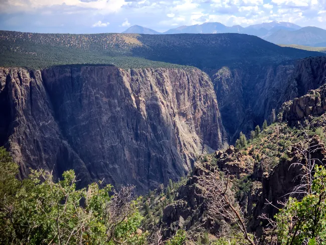 Dramatic view of Black Canyon’s vertical cliffs rising from forested slopes, with hazy mountain peaks in the background.